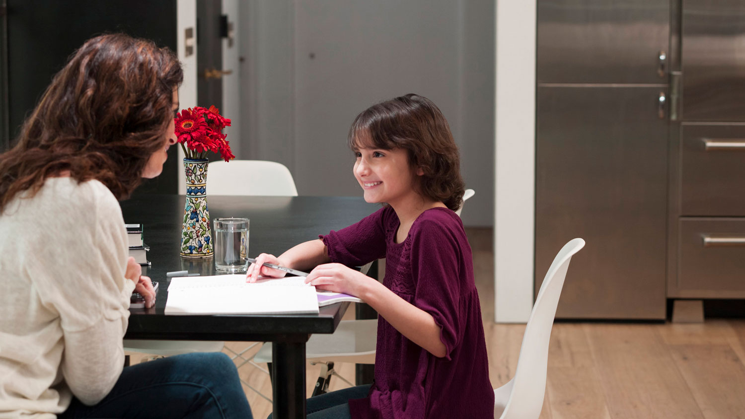 Smiling young girl sitting at a dining table doing homework with a woman across from her, in a modern kitchen with light wood floors and a vase of red flowers on the table.