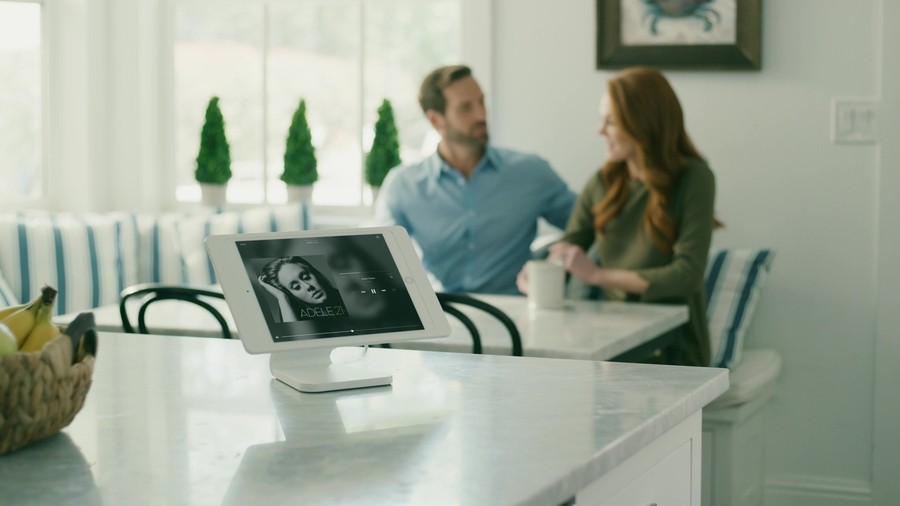 Two people sitting at a kitchen table. In the foreground is the Savant touch screen with an image of Adele. 