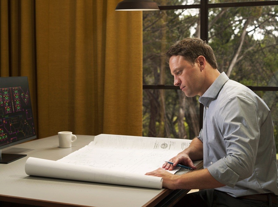 A man in a light blue dress shirt sits at a desk reviewing large architectural blueprints with a focused expression.