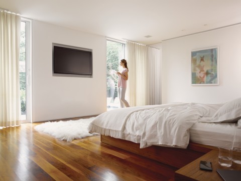 A woman standing by her bedroom window in a Savant home with in-ceiling speakers and a TV display.
