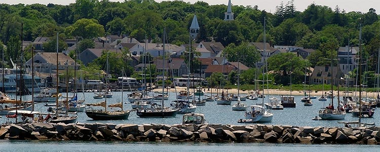 Harbor scene with numerous sailboats and small yachts anchored behind a rocky breakwater, with a quaint coastal town and church steeples nestled among trees in the background.