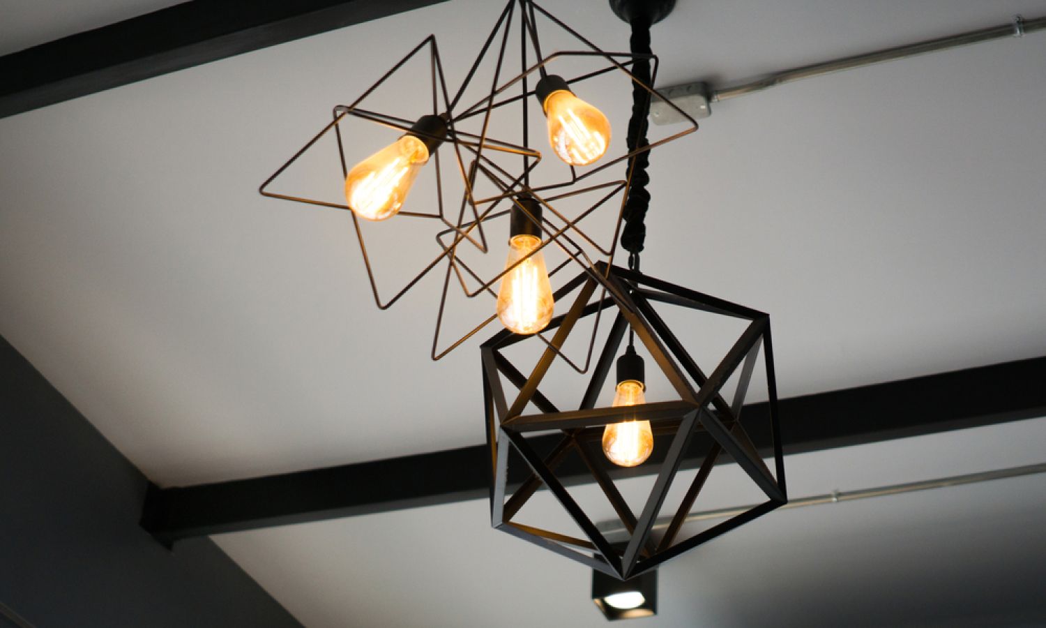 Close-up of contemporary ceiling light fixture: exposed Edison bulbs in black geometric wire and cube frames against a white ceiling.