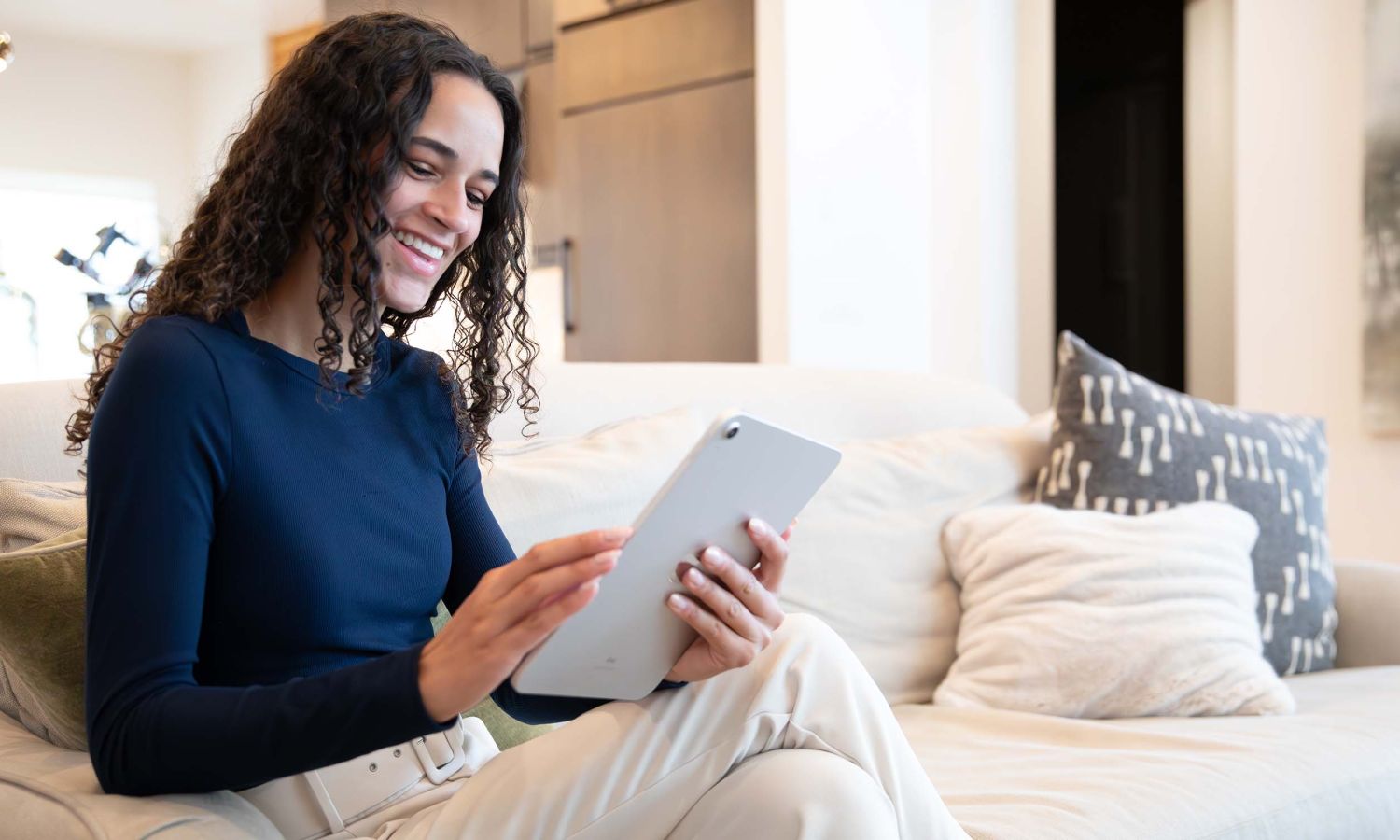 Person sitting on a light‑colored sofa using a tablet in a bright modern living room with decorative pillows and pendant lights.