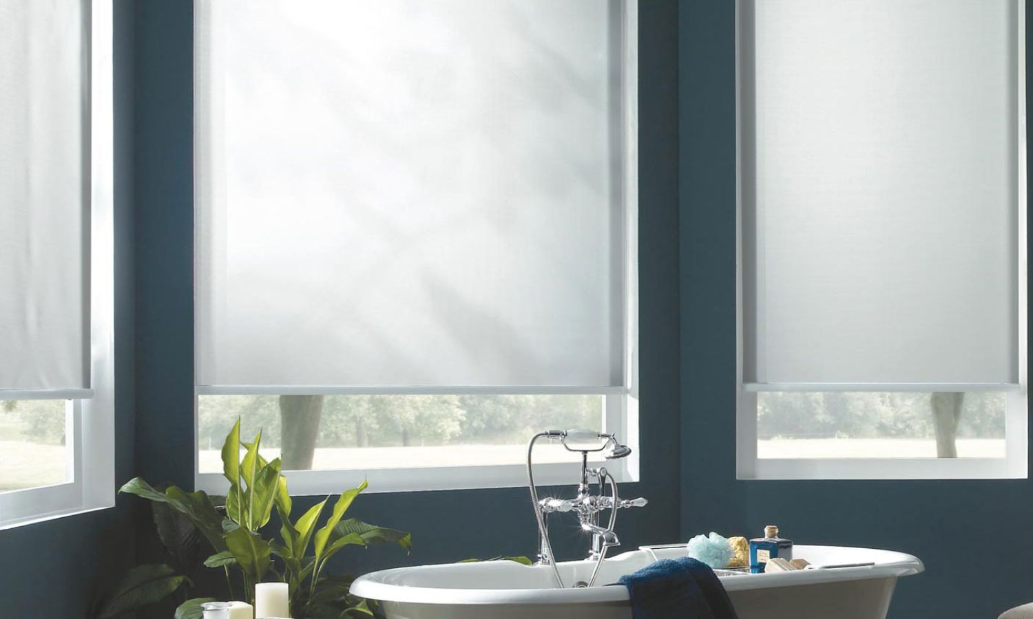 Modern bathroom with teal walls, a freestanding white bathtub and leafy plant in front of three motorized white roller shades filtering daylight.