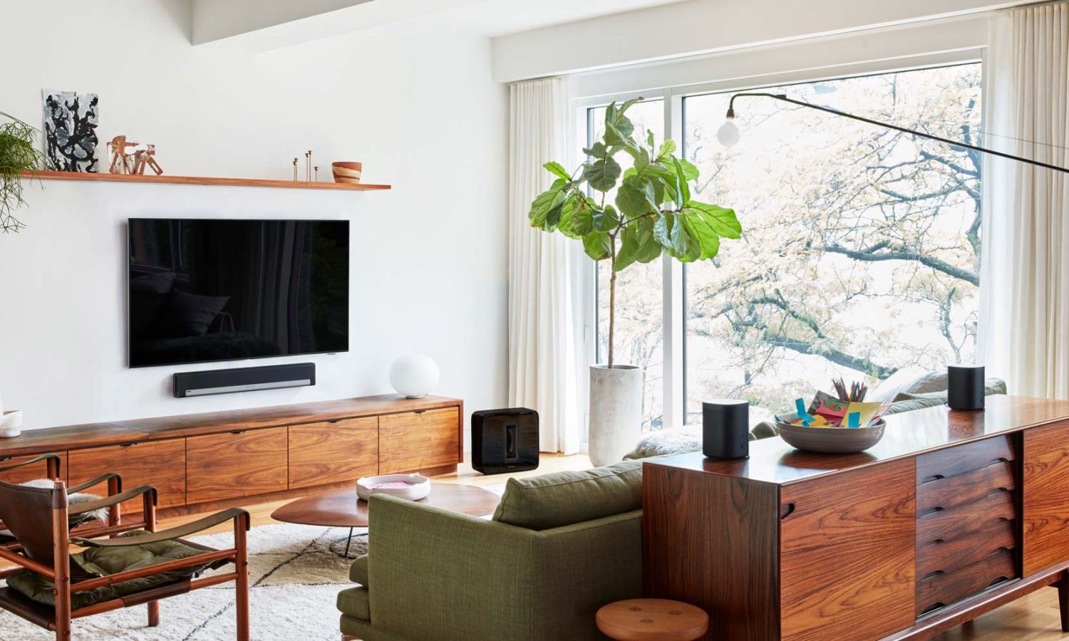 Modern living room with mid‑century wooden furniture, a wall‑mounted TV, a green sofa, and a large potted plant near tall windows.