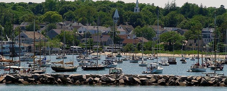 Harbor scene with numerous sailboats and small yachts anchored behind a rocky breakwater, with a quaint coastal town and church steeples nestled among trees in the background.