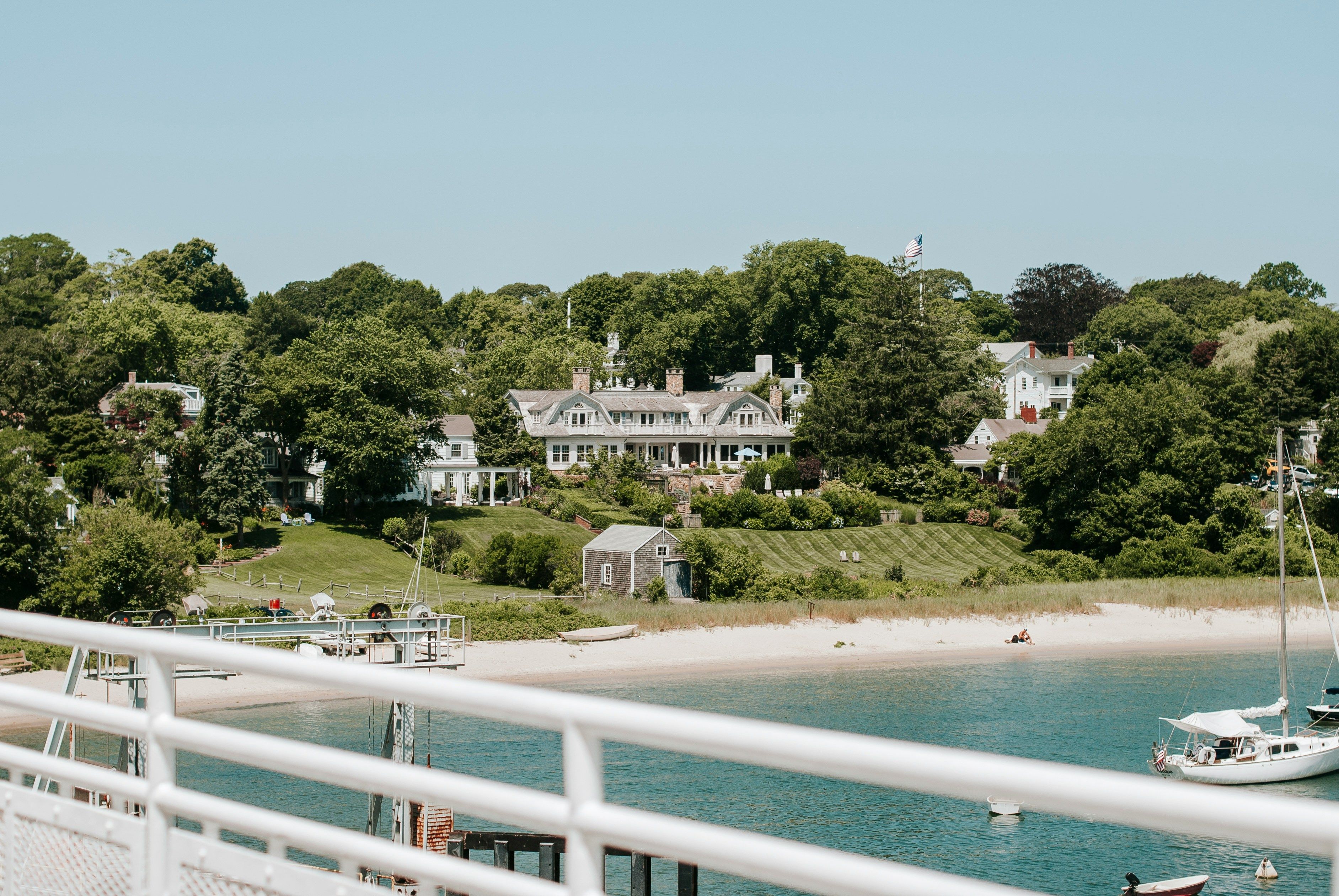 View from a boat or dock looking toward a New England–style coastal village, with large waterfront homes and lush trees above a sandy beach, sailboats anchored in the calm blue water, and white railings in the foreground.