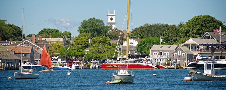 Harbor view of a coastal town with sailboats and motorboats on the water, shingled waterfront buildings along the shore, and a white clock tower rising above green trees in the background.